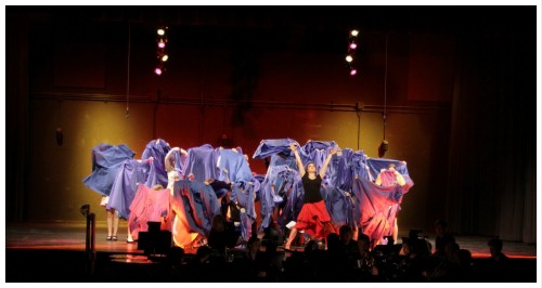 (Image: The Cast Holds Graduation Gowns
        in Front of Their Faces and Bodies while
        the Principal Dancer Stands with Her Arms
        Raised.)