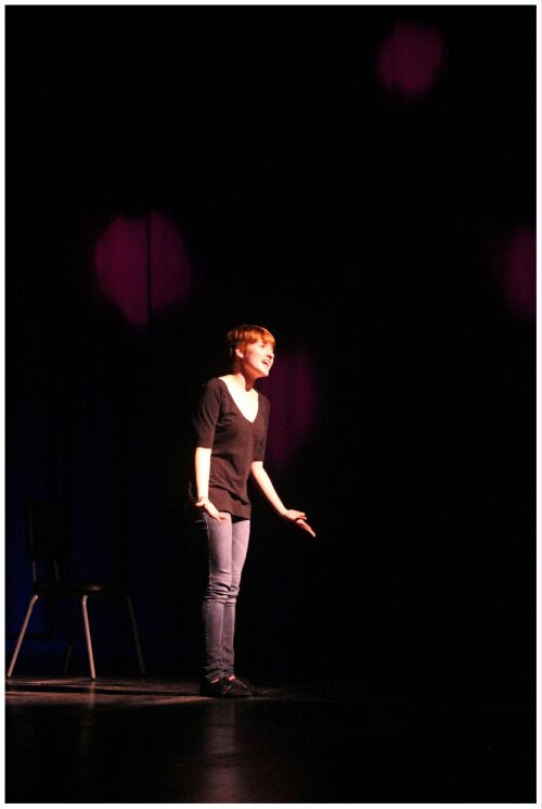 (Image: A Singer Stands on a Sparse Stage with Four
  Large Magenta Dots of Light on a Curtain Behind)